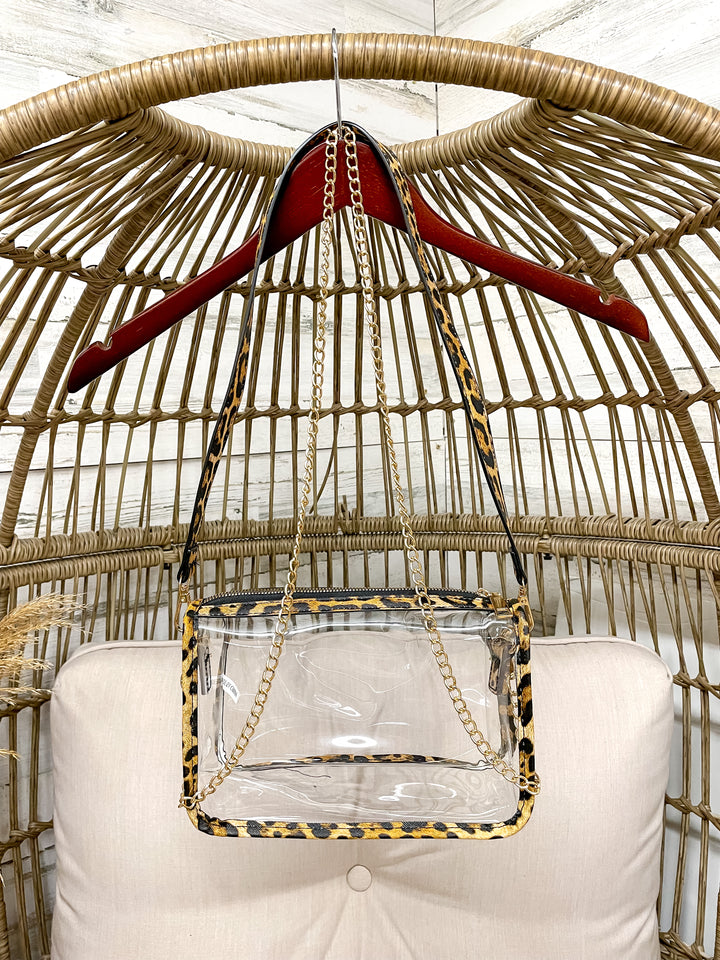 Clear handbag with leopard print trim hanging on a red hanger against a wicker chair background.