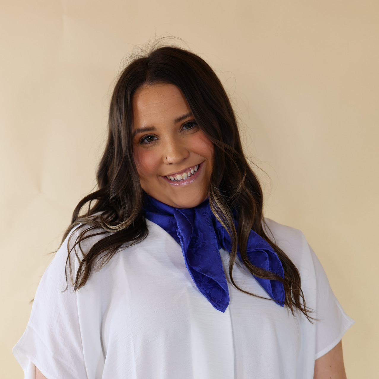 Brunette model is pictured wearing a white, Drop shoulder top with a solid blue scarf tied around her neck. Model is pictured in front of a beige background.
