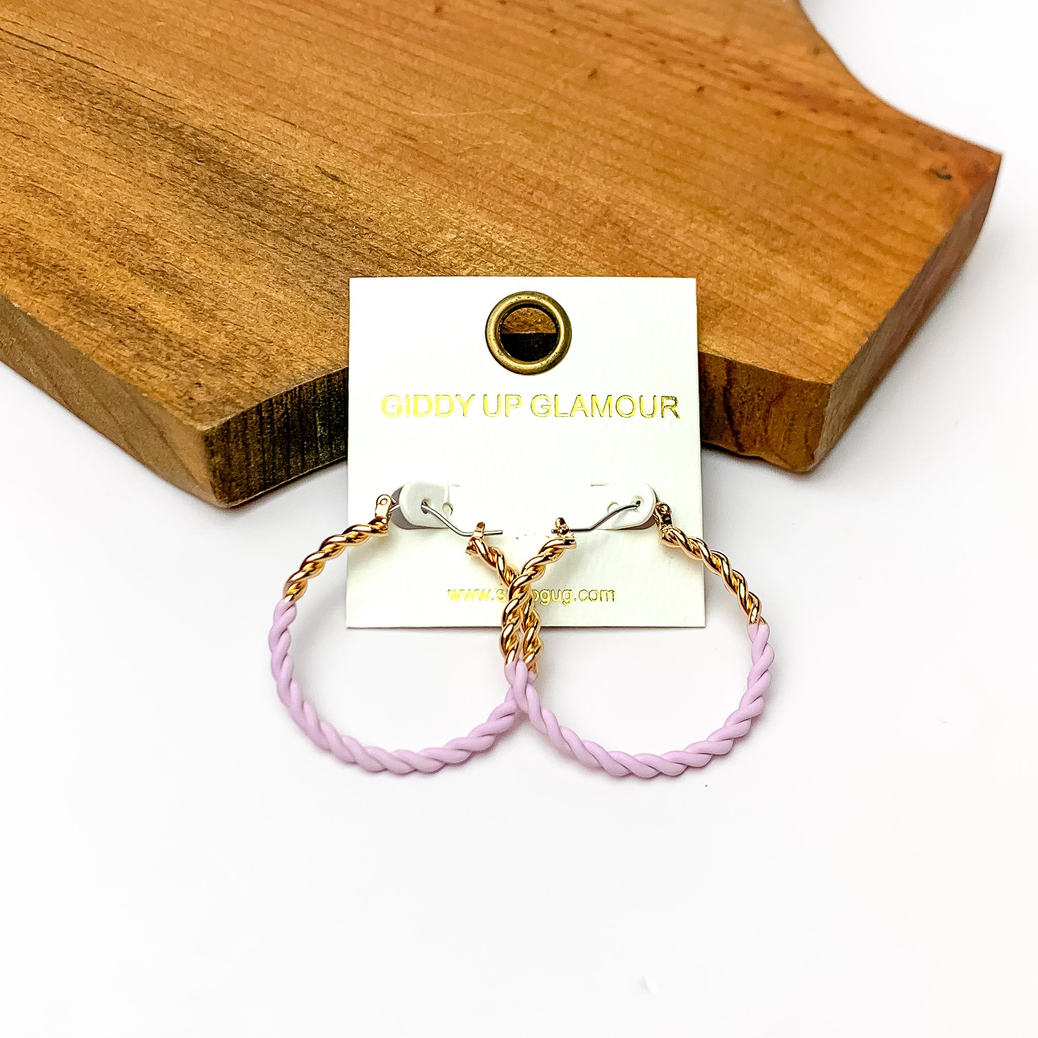 Twisted Gold Tone Hoop Earrings in Lavender. Pictured on a white background with a wood piece behind it.