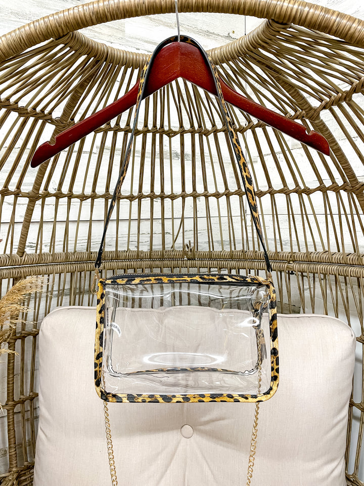 Clear handbag with leopard print accents on a red hanger against a wicker chair background