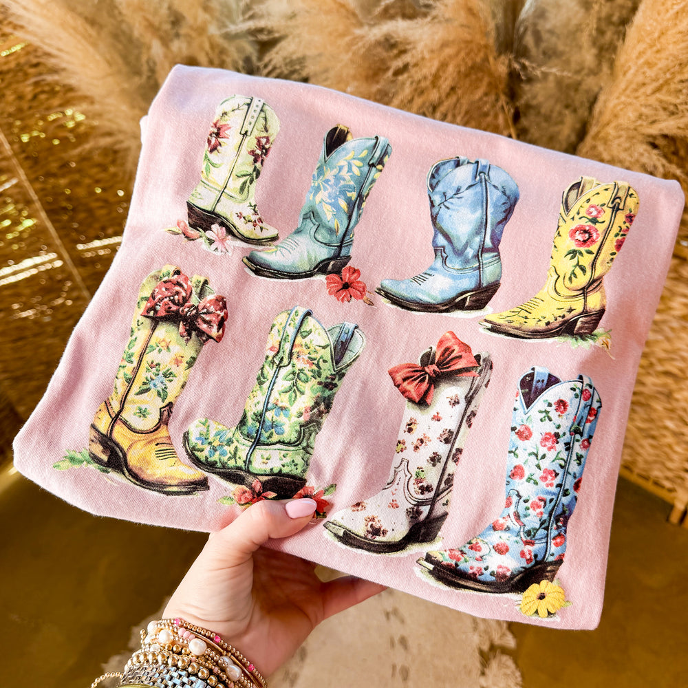 Pale blush pink tee printed with a grid of floral patterned cowboy boots and bows, held in hand.