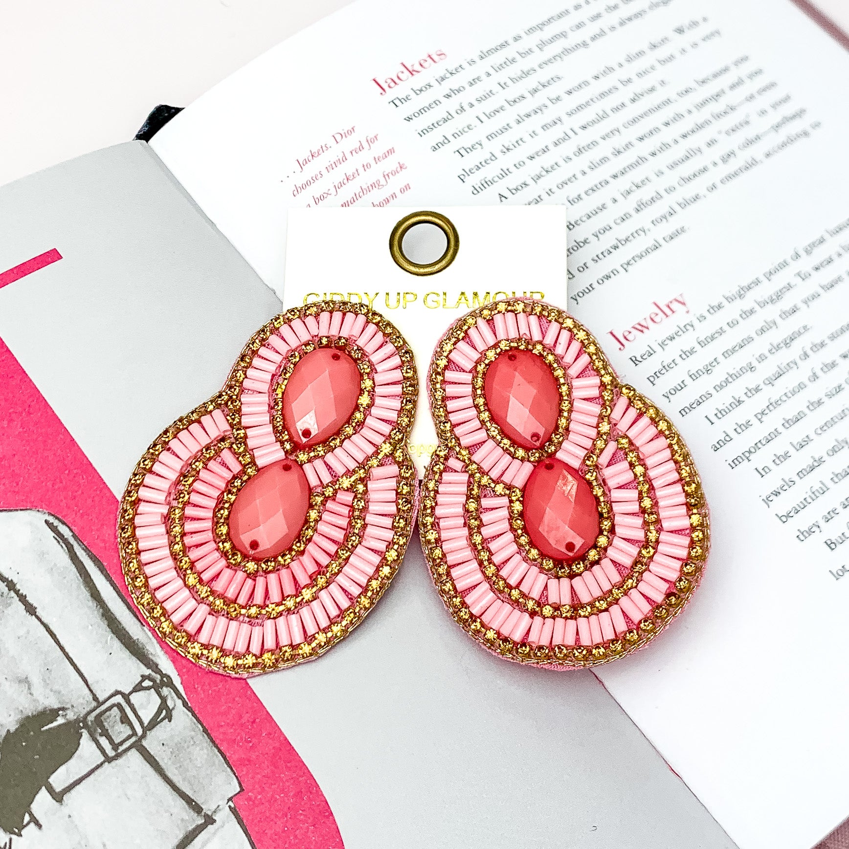 Dazzling Oval Earrings Outlined in Gold Tone Crystals with Beads in Light Pink. Pictured on a white background with a book opened behind the earrings.