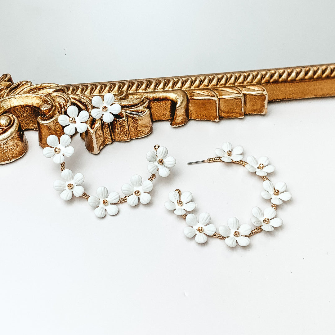 Gold hoop earrings with white flower charms outlining the earrings. These flowers have a gold bead center. One hoop is pictured laying against a gold mirror while the other hoop is laying flat on the white background. 