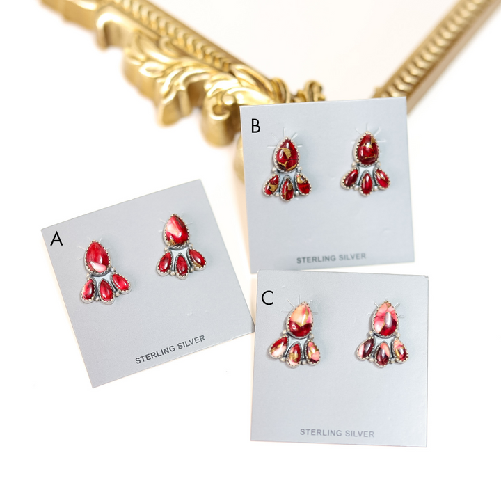 Three pairs of sterling silver Rose Dahlia stud earrings with deep red stones arranged in a floral cluster, displayed on earring cards near a decorative gold mirror frame.