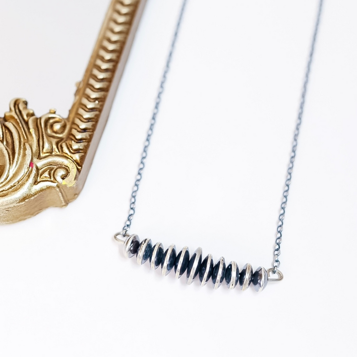 Close-up of Navajo pearl bar necklace with graduated sterling silver saucer beads on a delicate chain, laid flat next to a gold ornate frame.