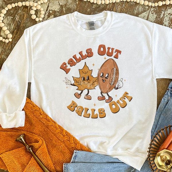 This white sweatshirt includes a crew neckline, long sleeves, and a cute hand drawn design of an orange leaf and football with the words "Fall Out" above them and "Balls Out" below them. The fonts are in rust orange and mustard yellow, perfect for Fall. This is shown as a flat lay with rolled sleeves and paired with a light wash pair of denim jeans. 