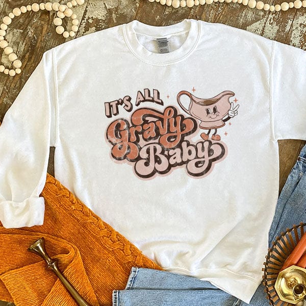This white sweatshirt includes a crew neckline, long sleeves, and a cute hand drawn design of a gravy boat holding up a peace sign with its fingers and the words "It's All Gravy Baby" in light peach and orange colors and a mix of fun fonts. This is shown as a flat lay with rolled sleeves and paired with light wash denim jeans. 
