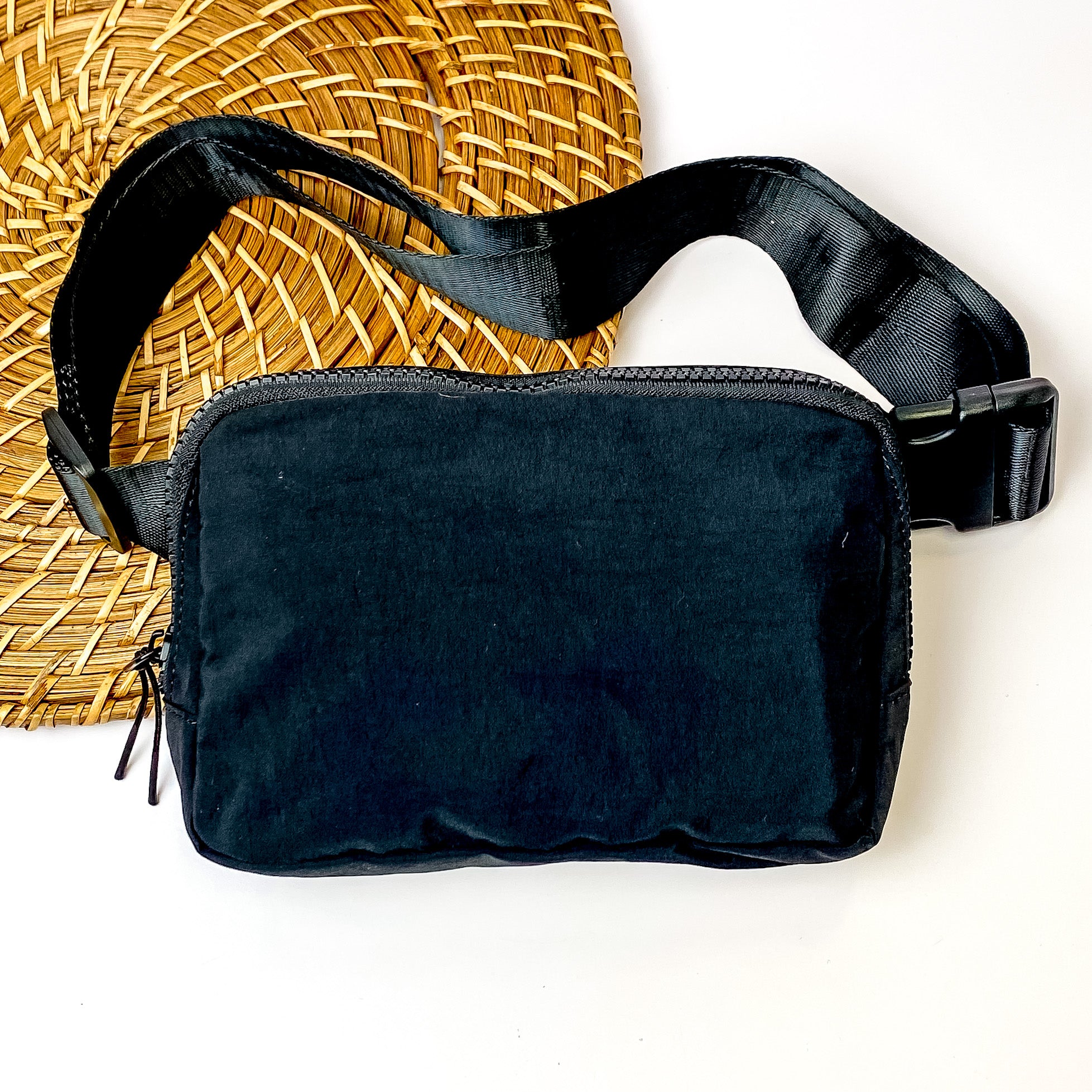 Pictured is a rectangle fanny pack with a top zipper with tassel in black. This bag also includes a black strap and black accents. This bag is pictured on a white and brown patterned background. 