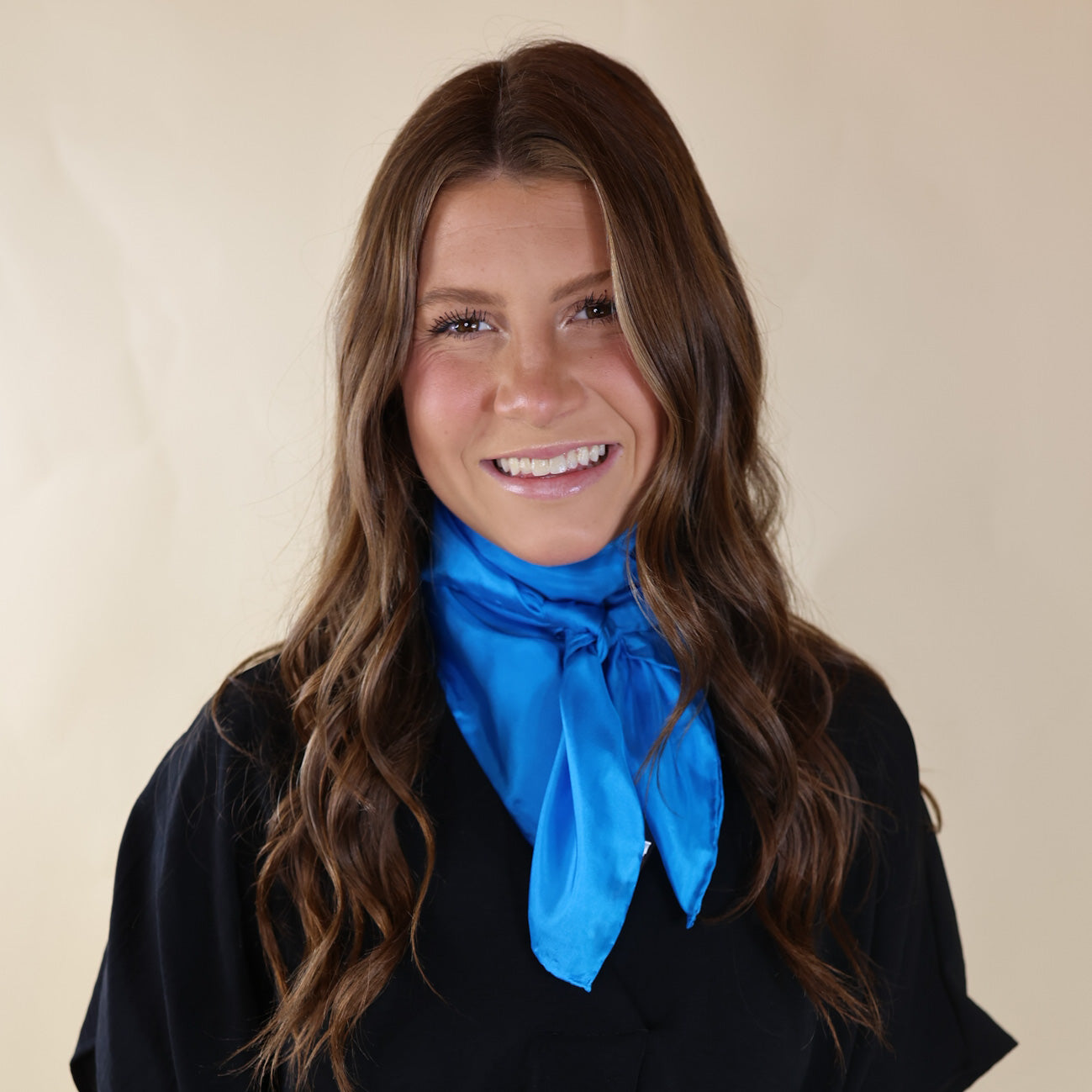 Brunette model is pictured wearing a Black, Drop shoulder top with a Solid Blue scarf tied around her neck. Model is pictured in front of a beige background. 
