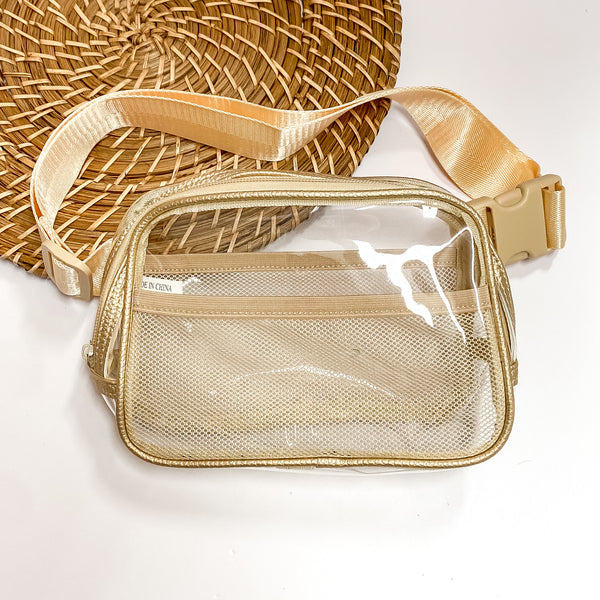 Pictured is a rectangle clear fanny pack with a gold outline. This bag also includes an ivory strap, ivory accents, and ivory mesh pockets. This bag is pictured on a white and brown patterned background. 