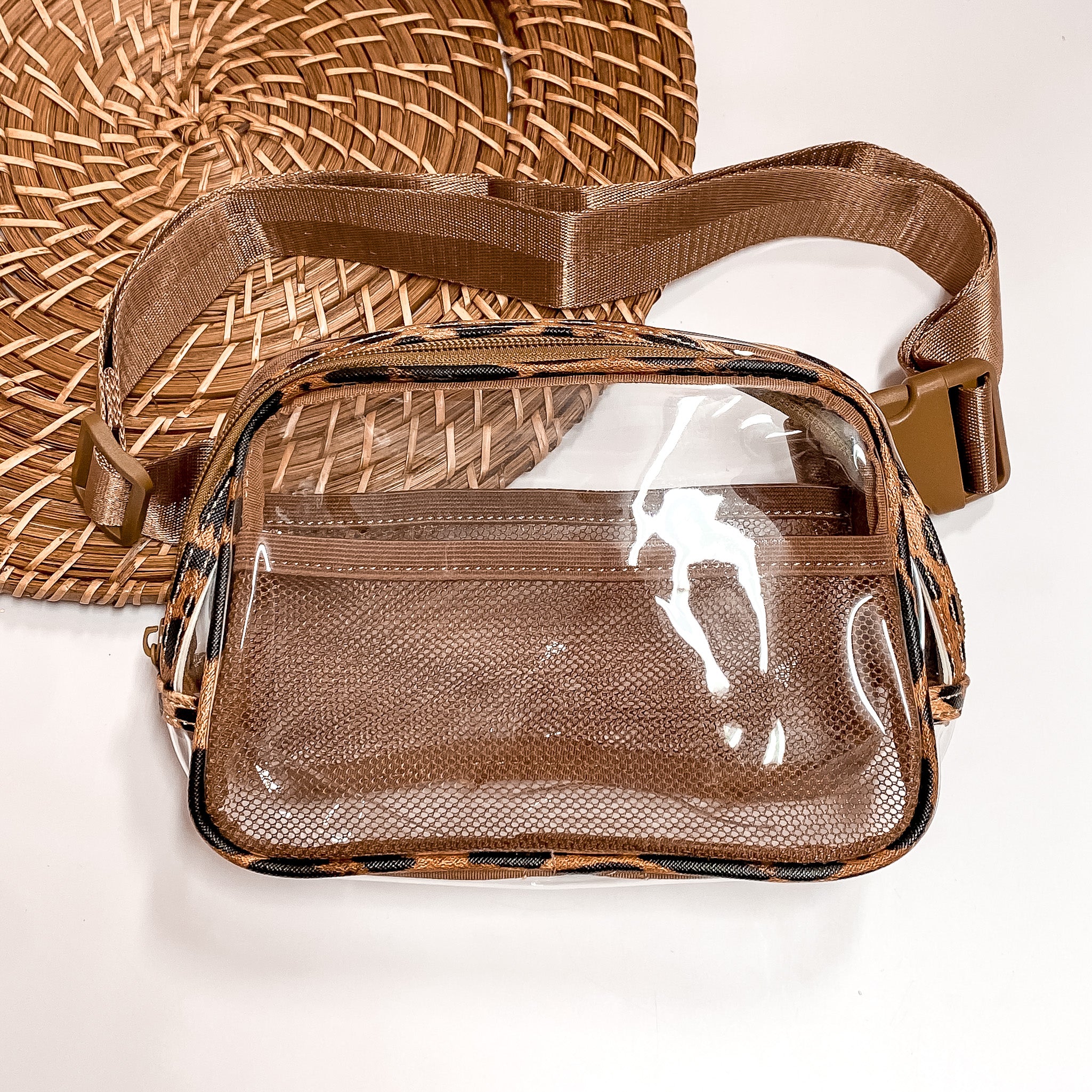 Pictured is a rectangle clear fanny pack with a leopard print outline. This bag also includes a tan strap, tan accents, and tan mesh pockets. This bag is pictured on a white and brown patterned background. 