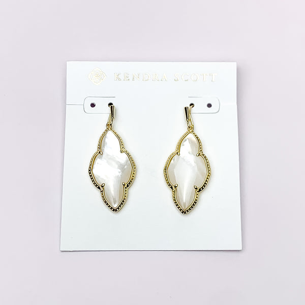 Gold drop earrings in a quatrefoil shape with ivory mother of pearl stone. These earrings are pictured on a white earring holder on a white background. 
