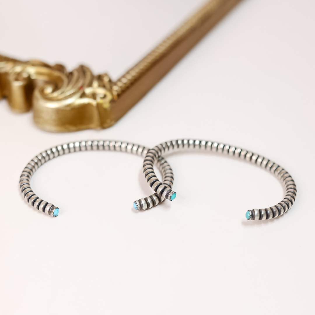 Adjustable sterling silver cuff bracelet with striped design and turquoise cabochons on the ends, styled on white background with gold mirror frame