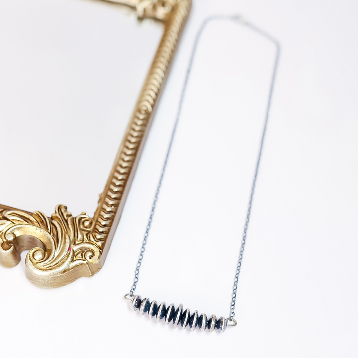 Close-up of Navajo pearl bar necklace with graduated sterling silver saucer beads on a delicate chain, laid flat next to a gold ornate frame.