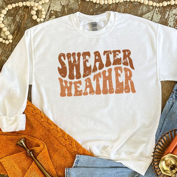 This white sweatshirt includes a crew neckline, long sleeves, and cute hand drawn design that says "Sweater Weather" in rust and orange heather uppercase bubble font. This sweatshirt is shown as a flat lay with rolled sleeves and a light wash pair of denim jeans. 
