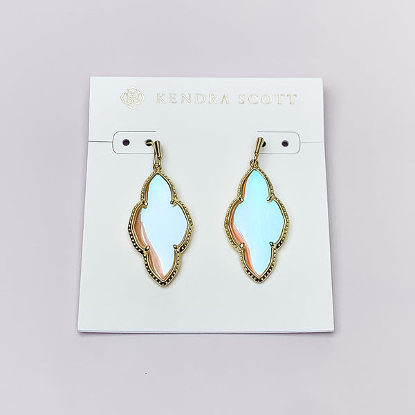 Gold drop earrings in a quatrefoil shape with dichroic glass. These earrings are pictured on a white earring holder on a white background. 