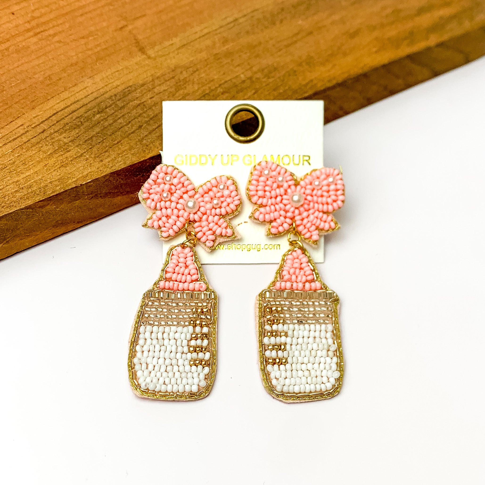 Baby Pink and White Beaded Bottle Earrings with Pink Bow Studs. Pictured on a white background with a wood piece at the top.