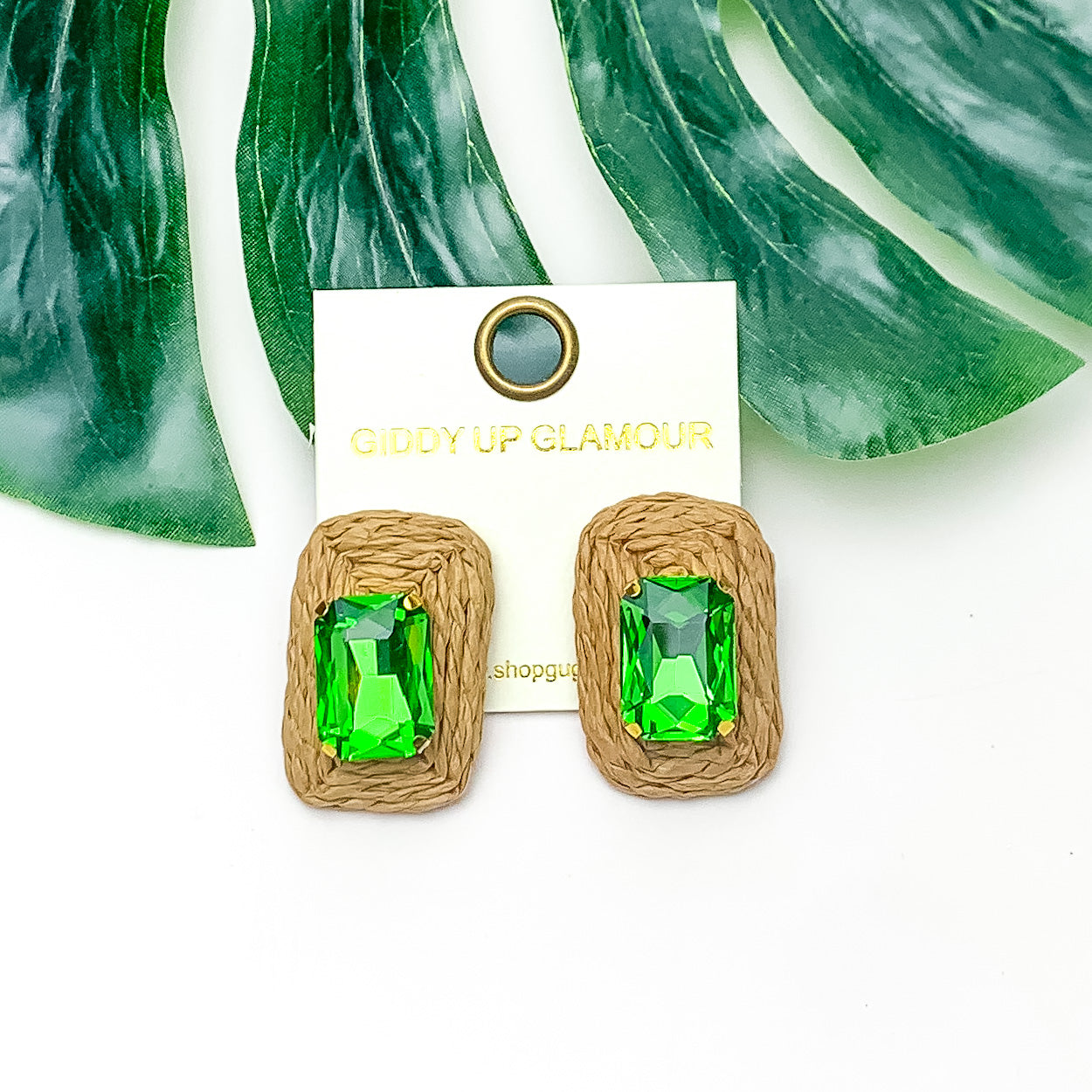 Truly Tropical Raffia Rectangle Earrings in Brown With Green Crystal. Pictured on a white background with the earrings laying on a large leaf.
