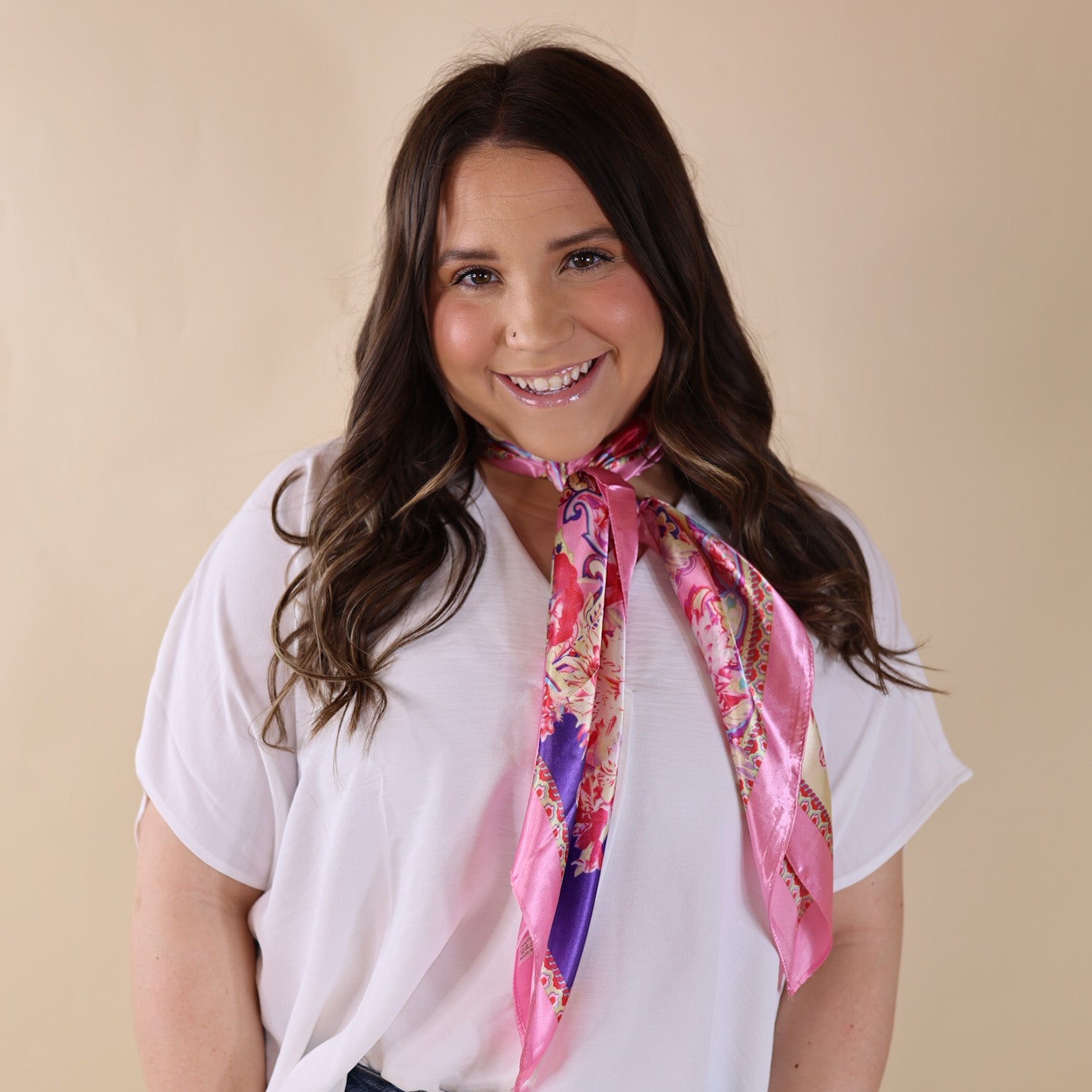 Brunette model is wearing a white drop shoulder top with a multicolored pink scarf wrapped and tied around her neck. She is pictured in front of a beige background.