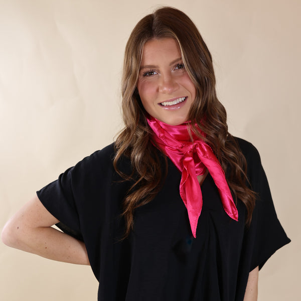 Brunette model is wearing a black drop shoulder top with a pink scarf wrapped and tied around her neck. She is pictured in front of a beige background.