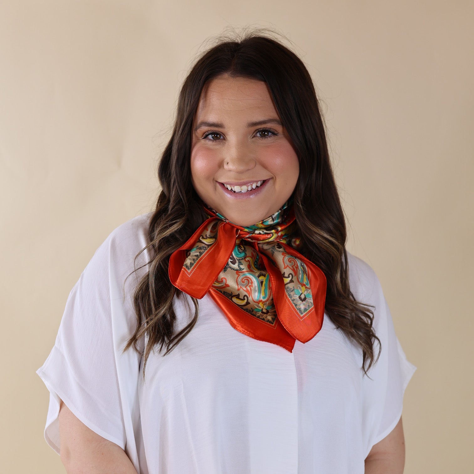 Brunette model is wearing a white drop shoulder top with a beige printed scarf with a red-orange border wrapped and tied around her neck. She is pictured in front of a beige background.