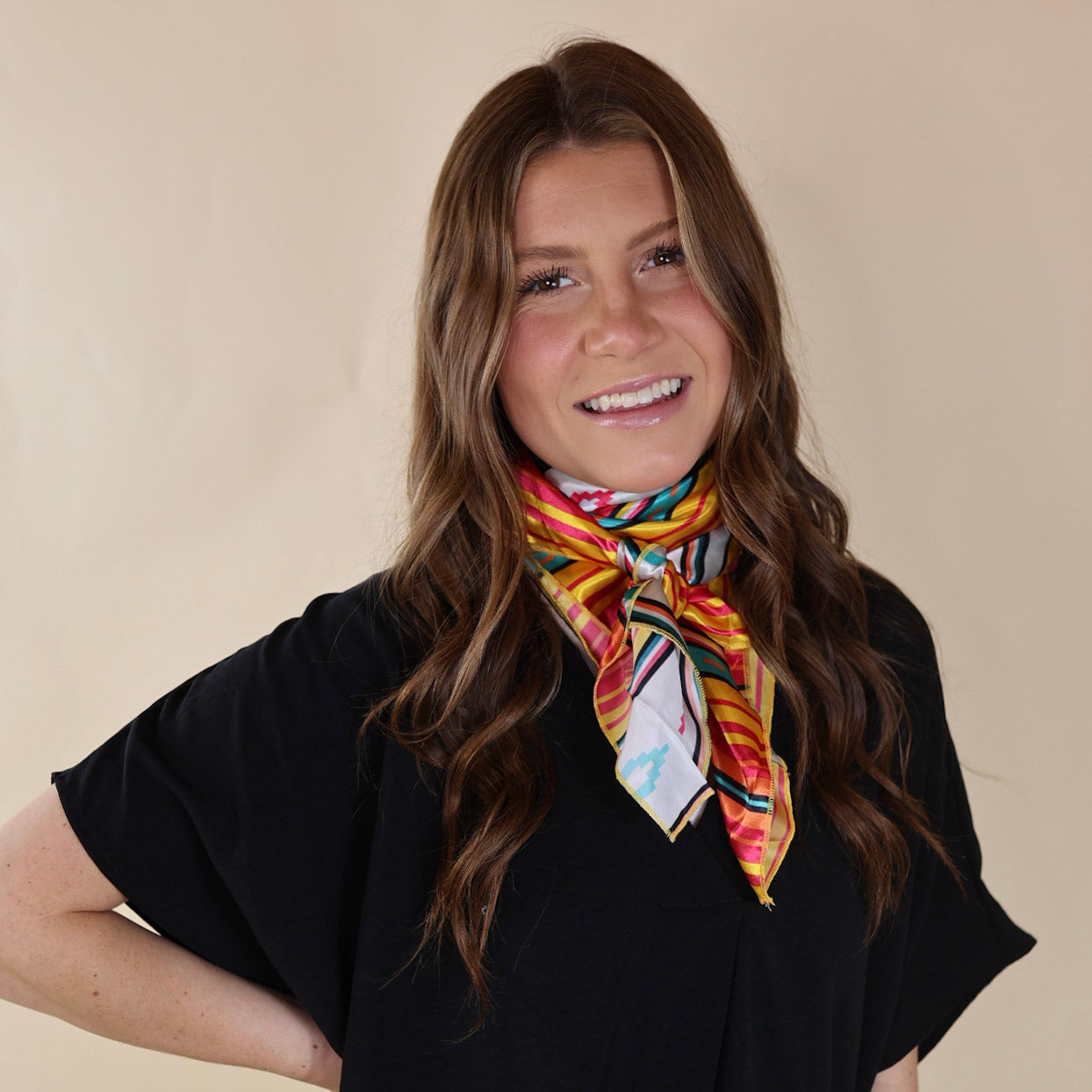 Brunette model wearing a black, drop shoulder top with a multicolored striped scarf tied around her neck. Model is pictured in front of a beige background. 