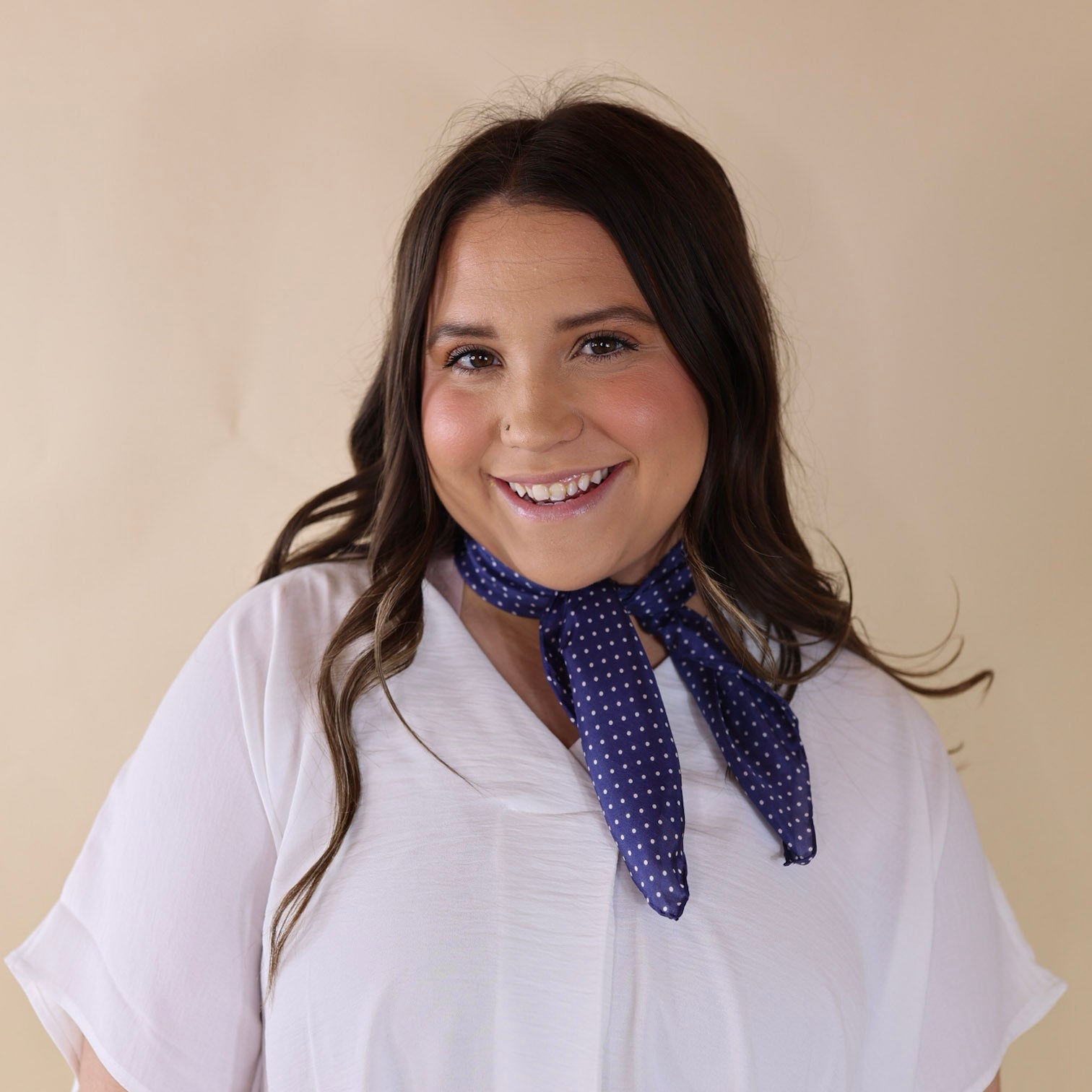 Brunette model wearing a white, off the shoulder top with a navy blue scarf with a white dotted print tied around her neck. Model is pictured in front of a beige background. 