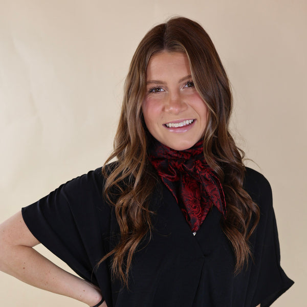 Brunette model wearing a black, off the shoulder top with a black and red baroque print scarf tied around her neck. Model is pictured in front of a beige background. 