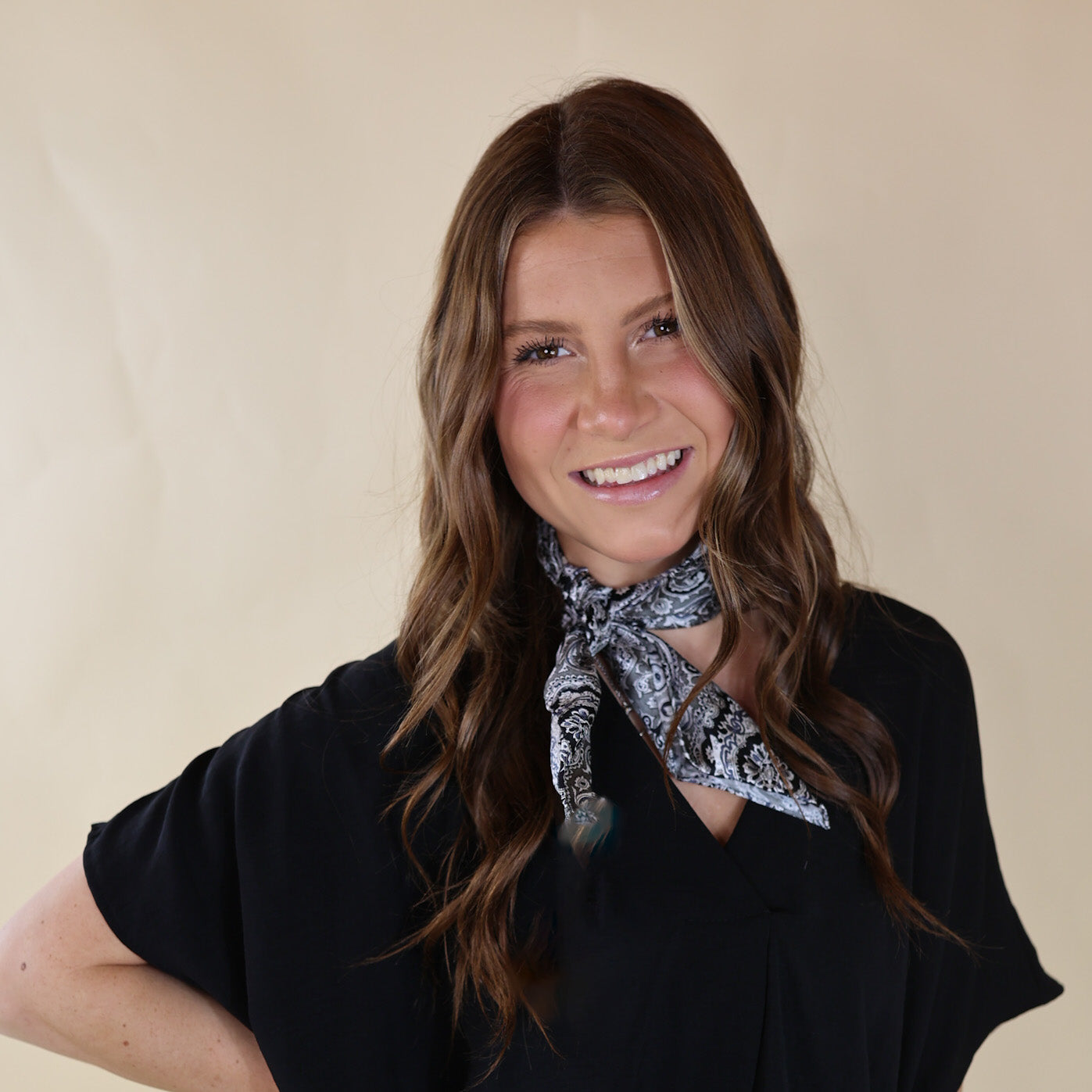 Brunette model wearing a black, drop shoulder top with Black and Silver scarf tied around her neck. Model is pictured in front of a beige background.