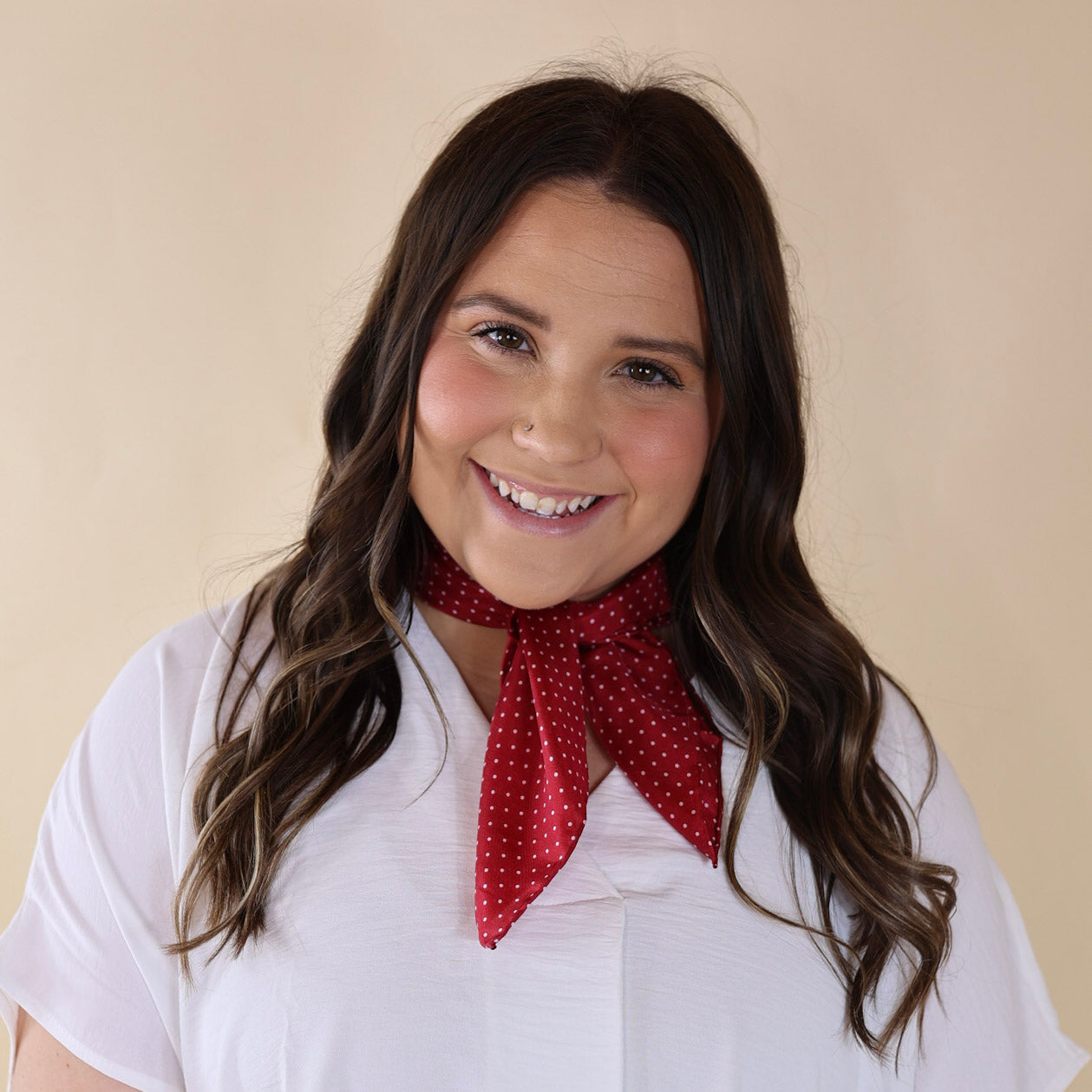 Brunette model wearing a white, off the shoulder top with a red scarf with a white dotted print tied around her neck. Model is pictured in front of a beige background. 