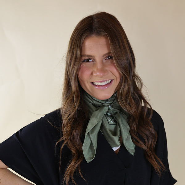 Brunette model wearing a short sleeve, black top with a sage green bandana scarf tied around her neck. This model is pictured in front of a beige background. 