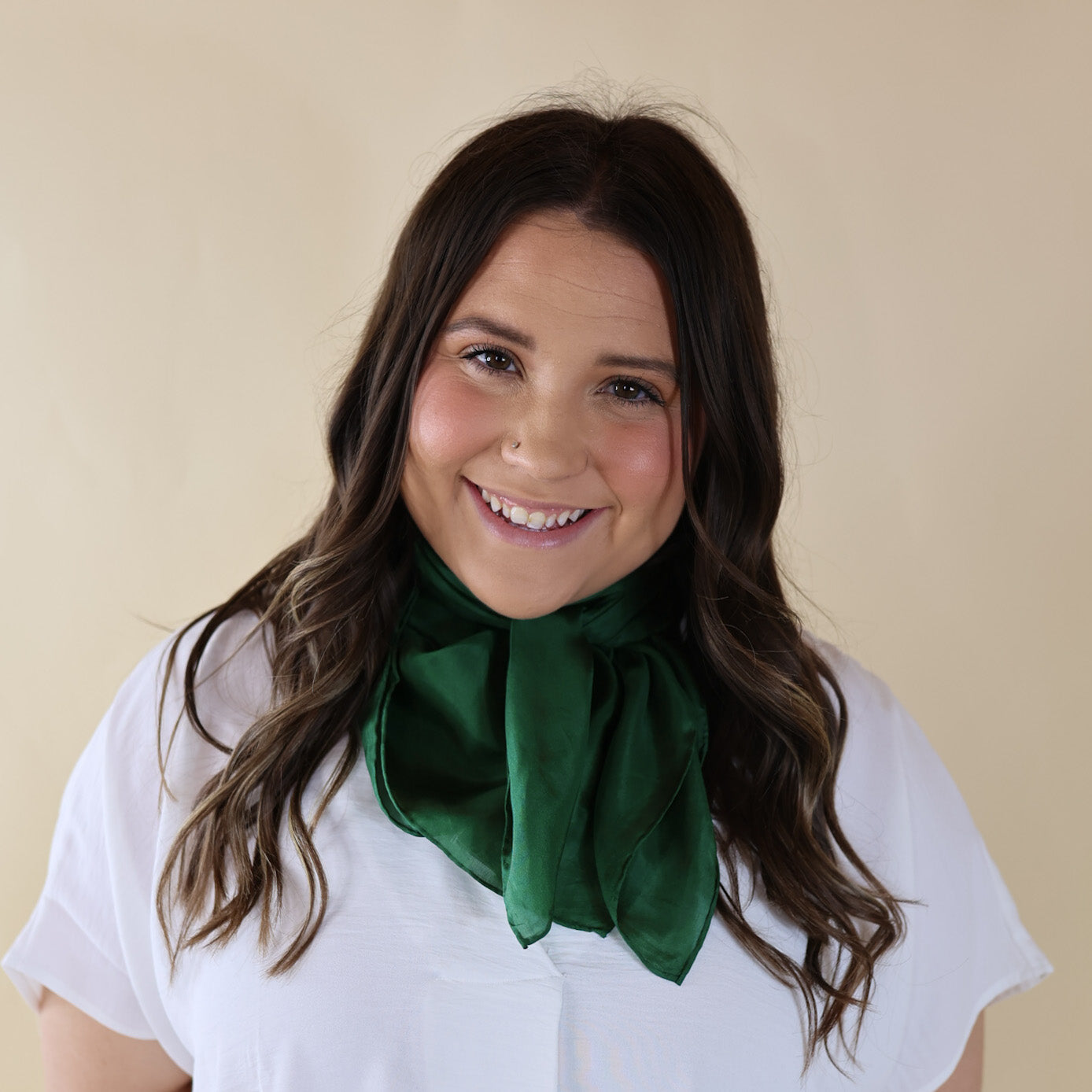 Brunette model wearing a short sleeve, white top with a eucalyptus green scarf tied around her neck. This model is pictured in front of a beige background. 