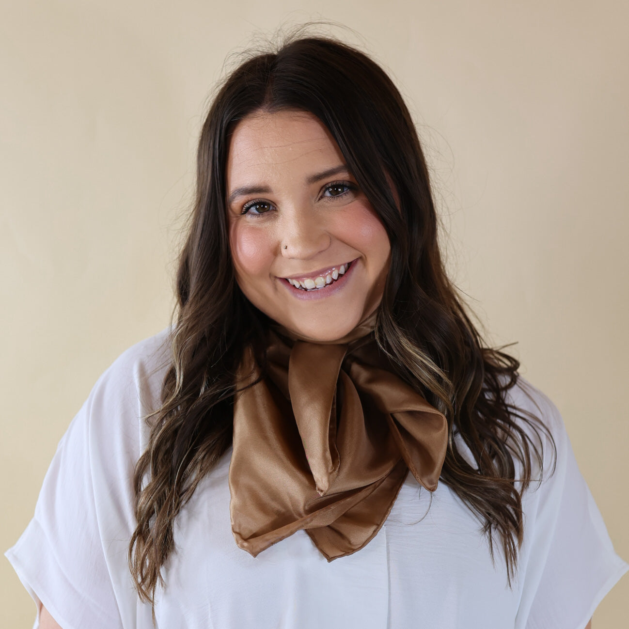 Brunette model wearing a short sleeve, white top with a tan scarf tied around her neck. This model is pictured in front of a beige background. 