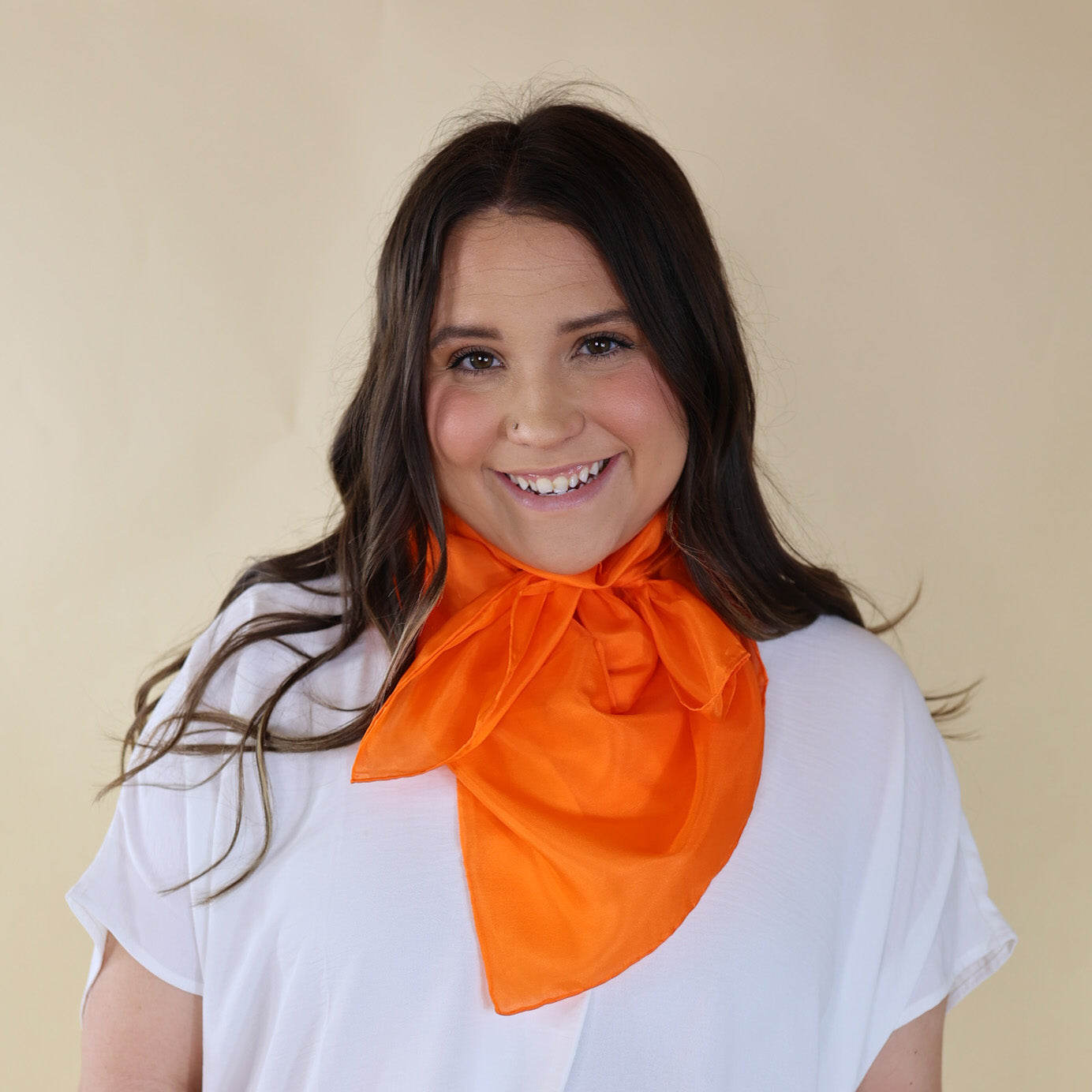 Brunette model wearing a white, drop shoulder top with bright orange scarf tied around her neck. Model is pictured in front of a beige background.