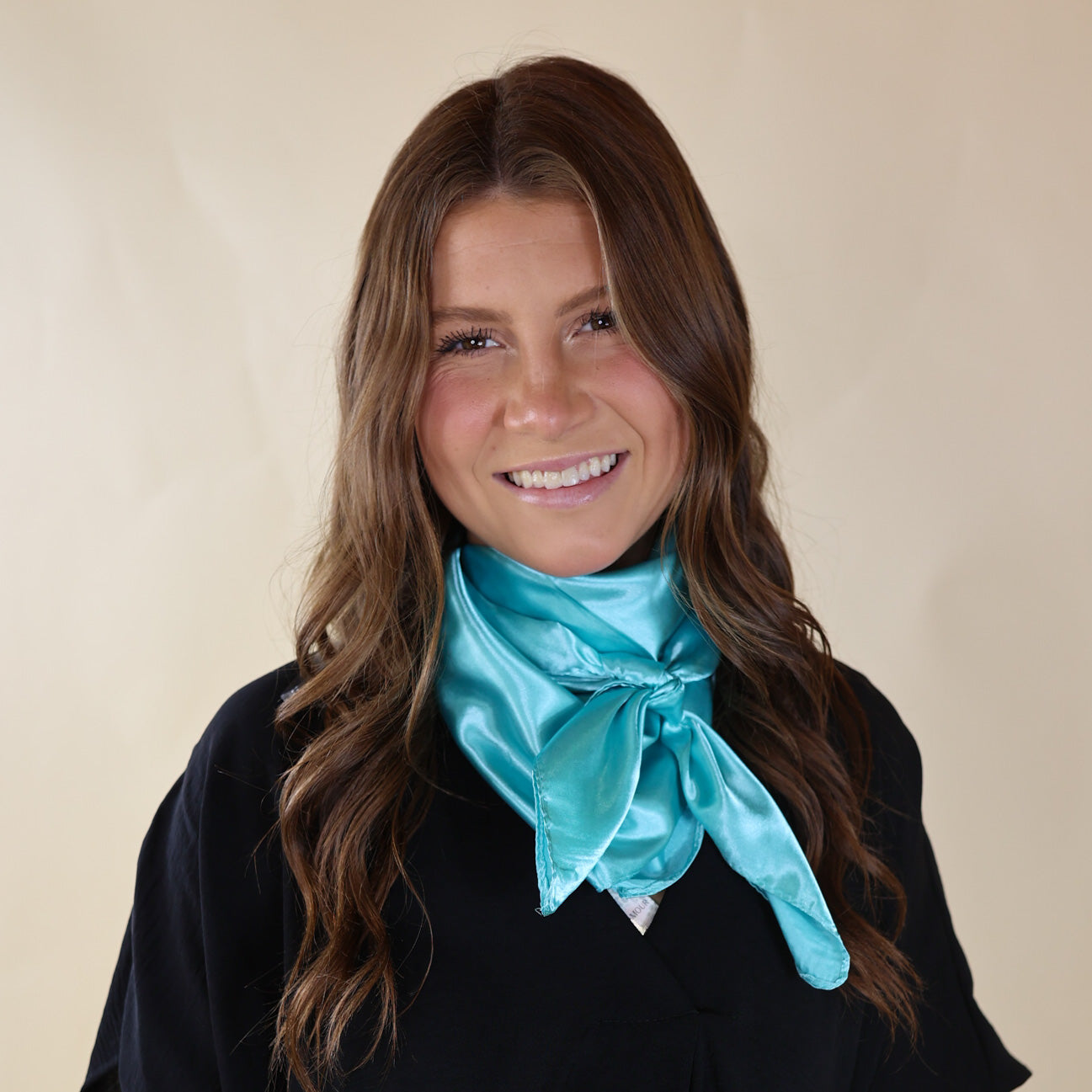 Brunette model wearing a short sleeve, black top with a light turquoise scarf tied around her neck. This model is pictured in front of a beige background. 