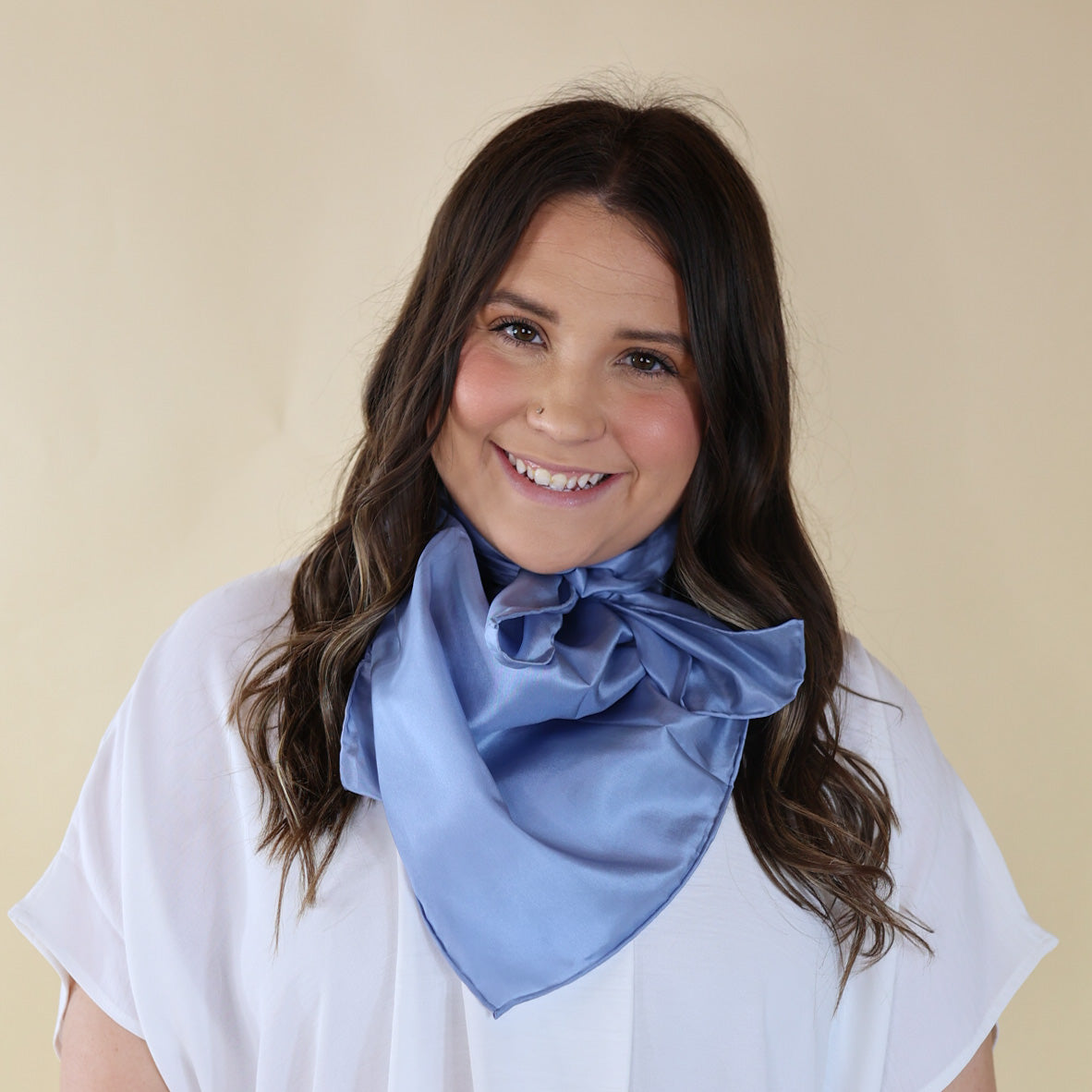 Brunette model wearing a short sleeve, white top with a slate blue scarf tied around her neck. This model is pictured in front of a beige background. 