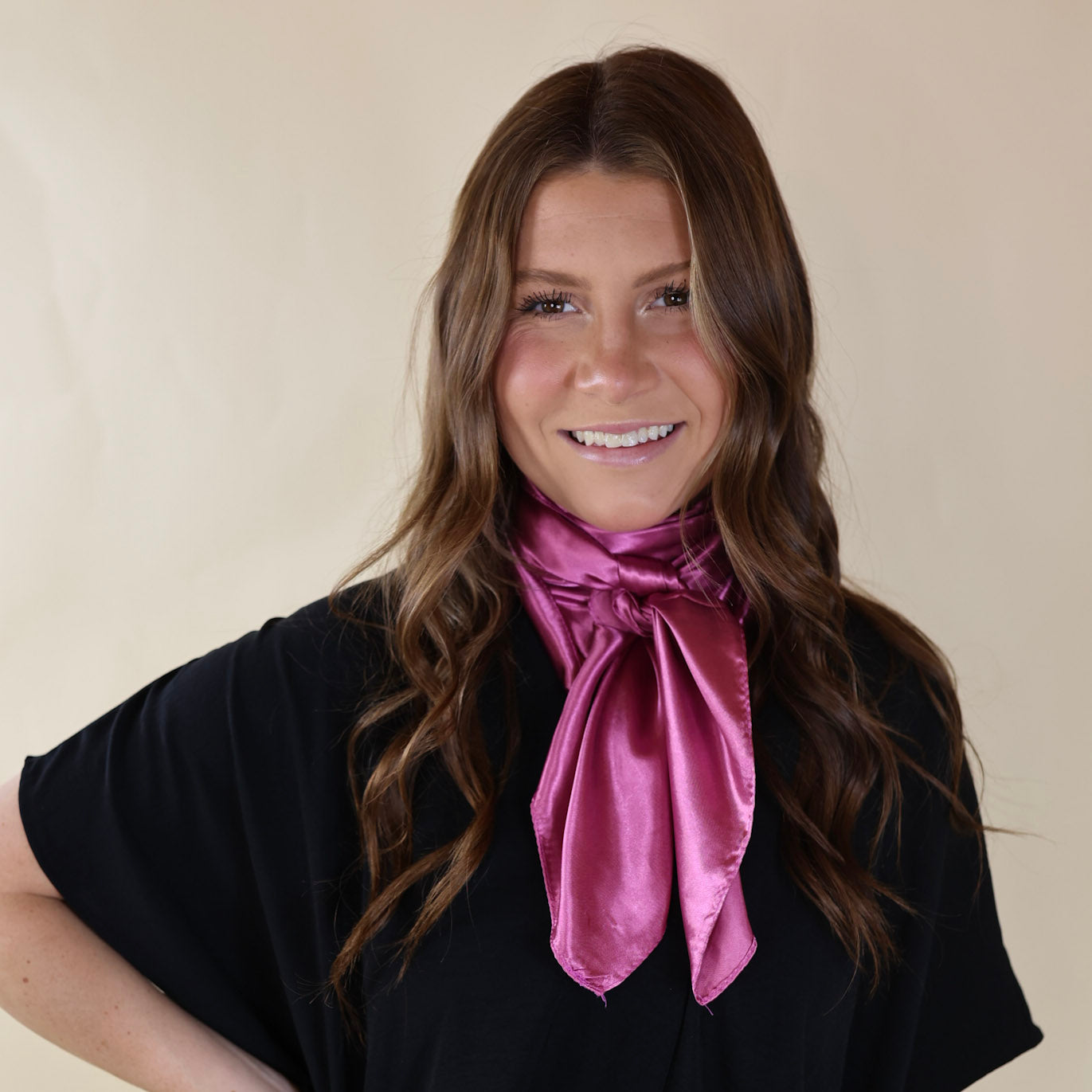 Brunette model wearing a black, drop shoulder top with a mauve pink scarf tied around her neck. Model is pictured in front of a beige background. 