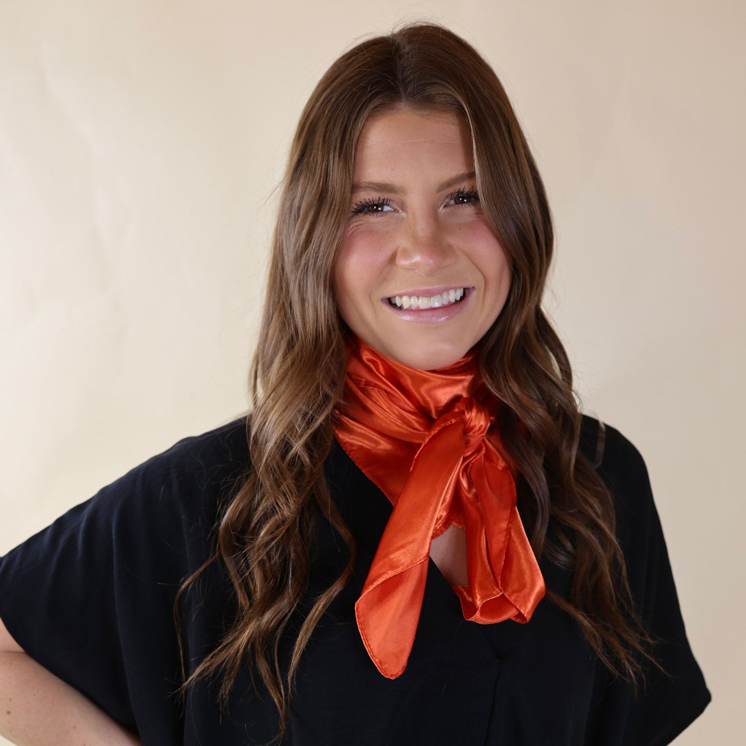 Brunette model wearing a black, drop shoulder top with a coral orange scarf tied around her neck. Model is pictured in front of a beige background. 