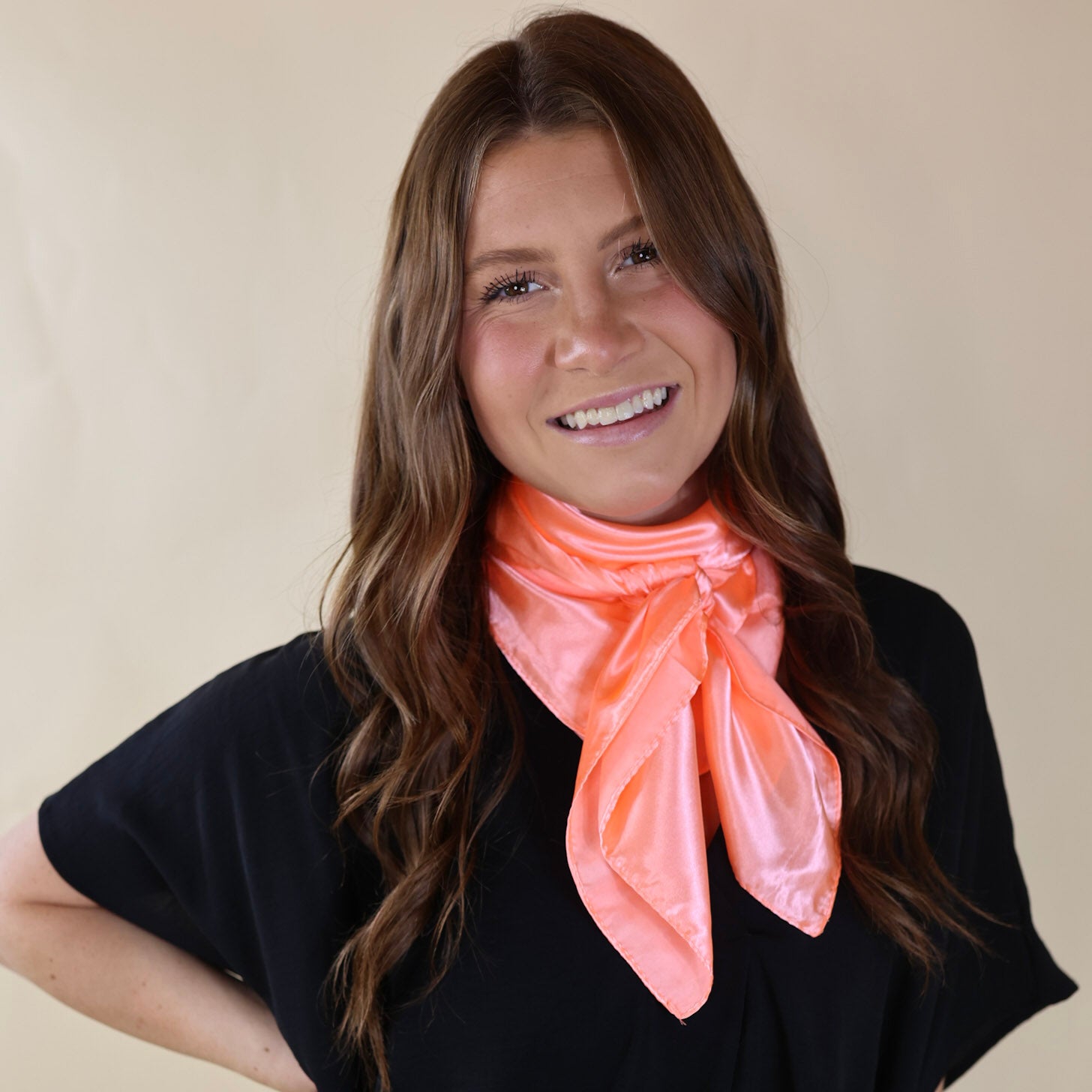 Brunette model wearing a black, drop shoulder top with a peach scarf tied around her neck. Model is pictured in front of a beige background. 