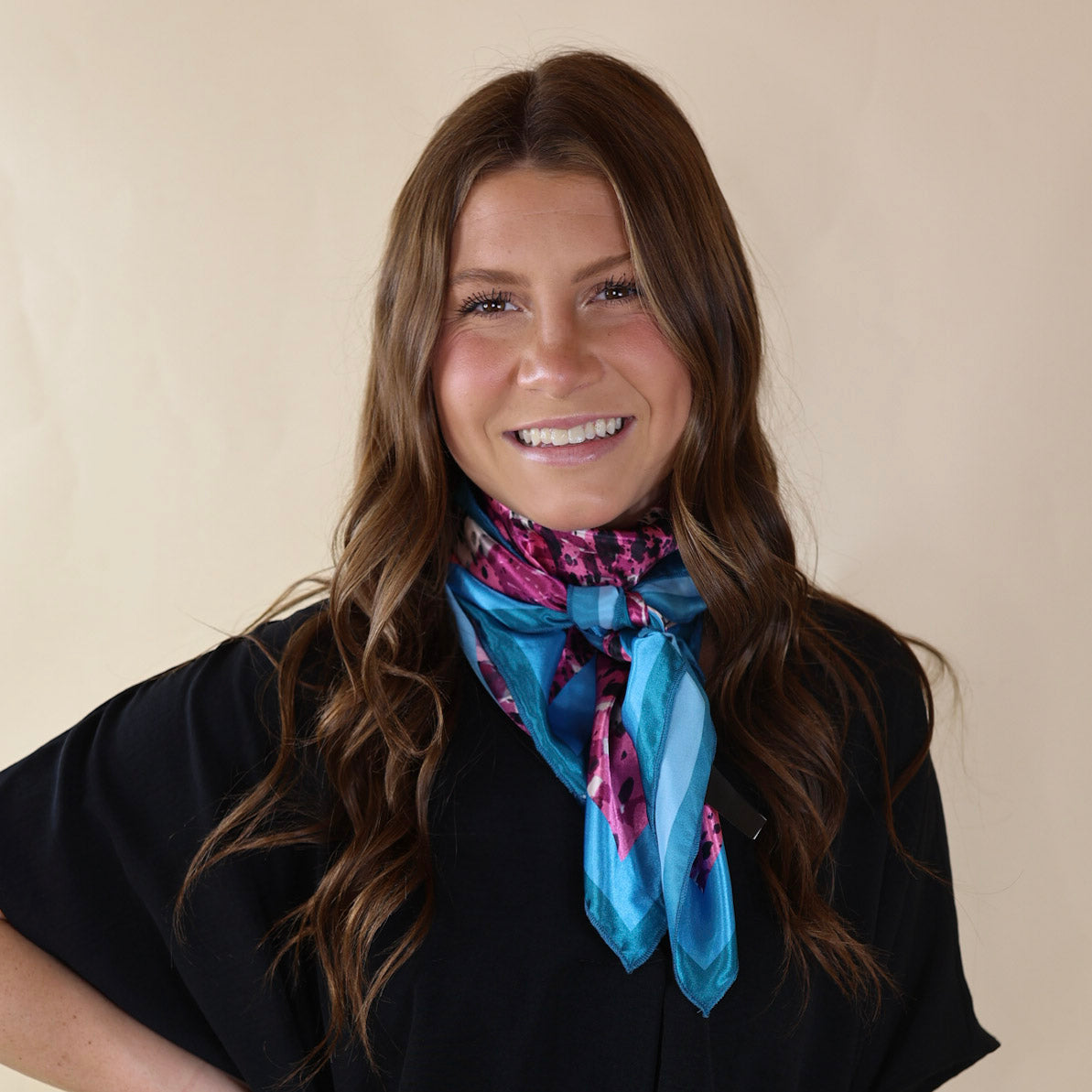 Brunette model wearing a black, drop shoulder top with a pink and beige cowprint and blue outline scarf with tied around her neck. Model is pictured in front of a beige background. 