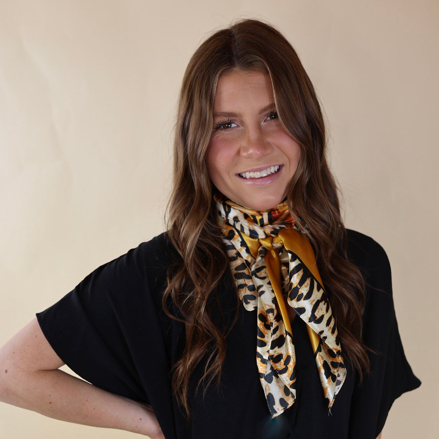 Brunette model wearing a black, drop shoulder top with mustard yellow and leopard outline scarf with tied around her neck. Model is pictured in front of a beige background. 