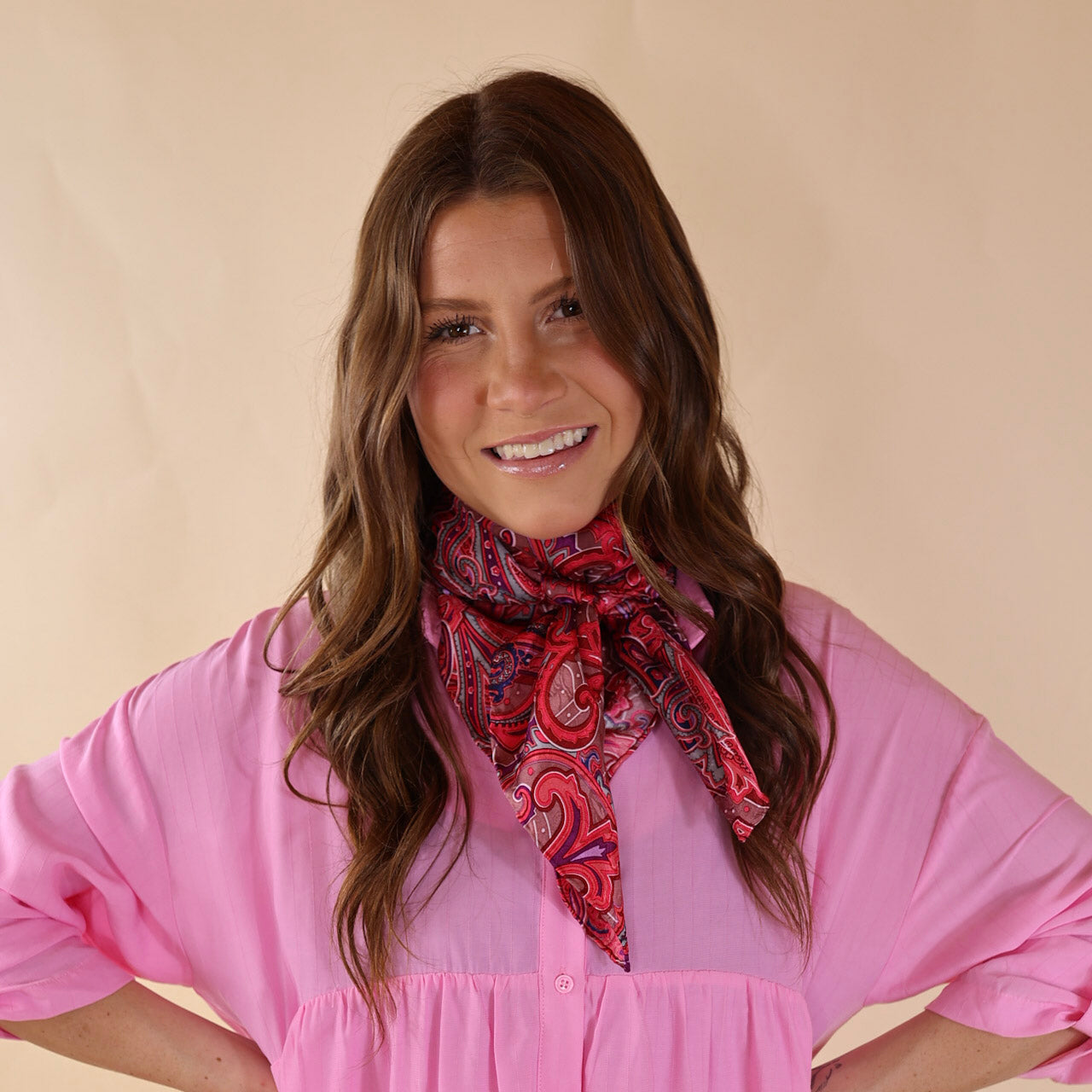 Brunette model wearing a pink, button up top with a paisly print scarf in a pink mix tied around her neck. Model is pictured in front of a beige background. 