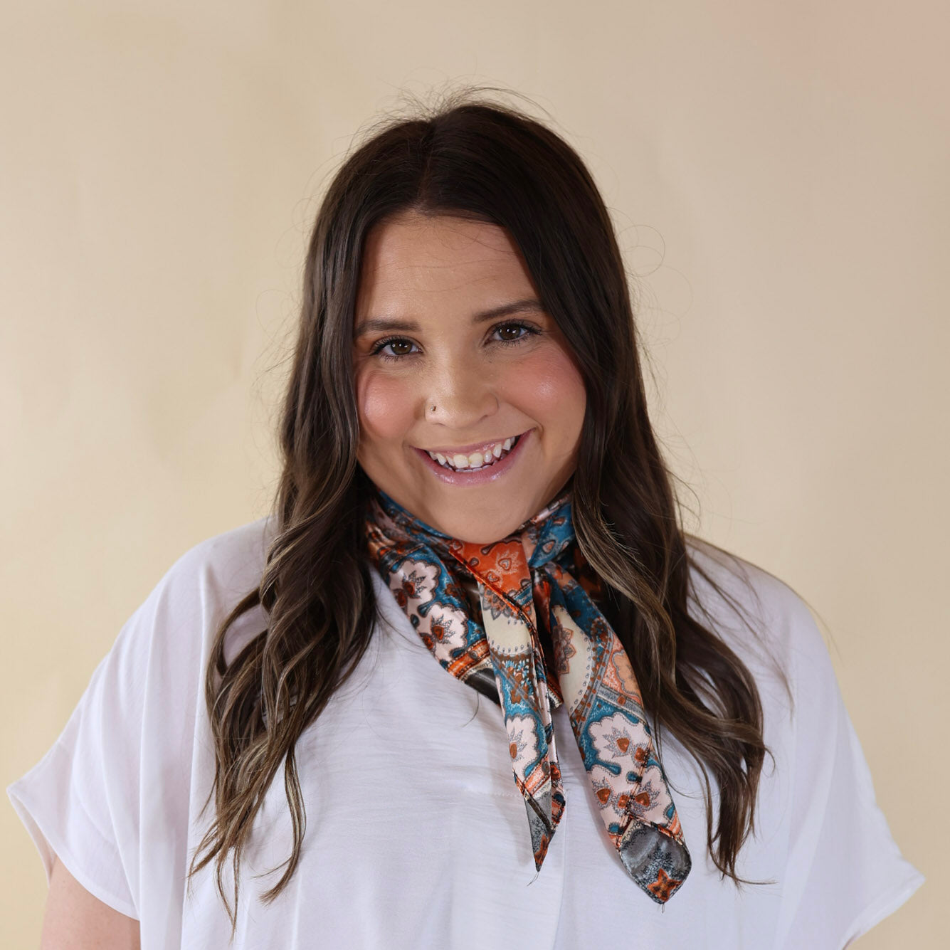 Brunette model wearing a white, drop shoulder top with multicolor paisley print scarf with tied around her neck. Model is pictured in front of a beige background. 