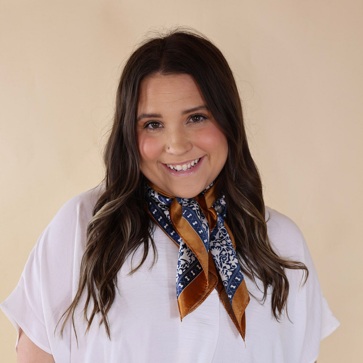 Brunette model wearing a white, drop shoulder top with blue, cream, and tan design scarf with tied around her neck. Model is pictured in front of a beige background. 
