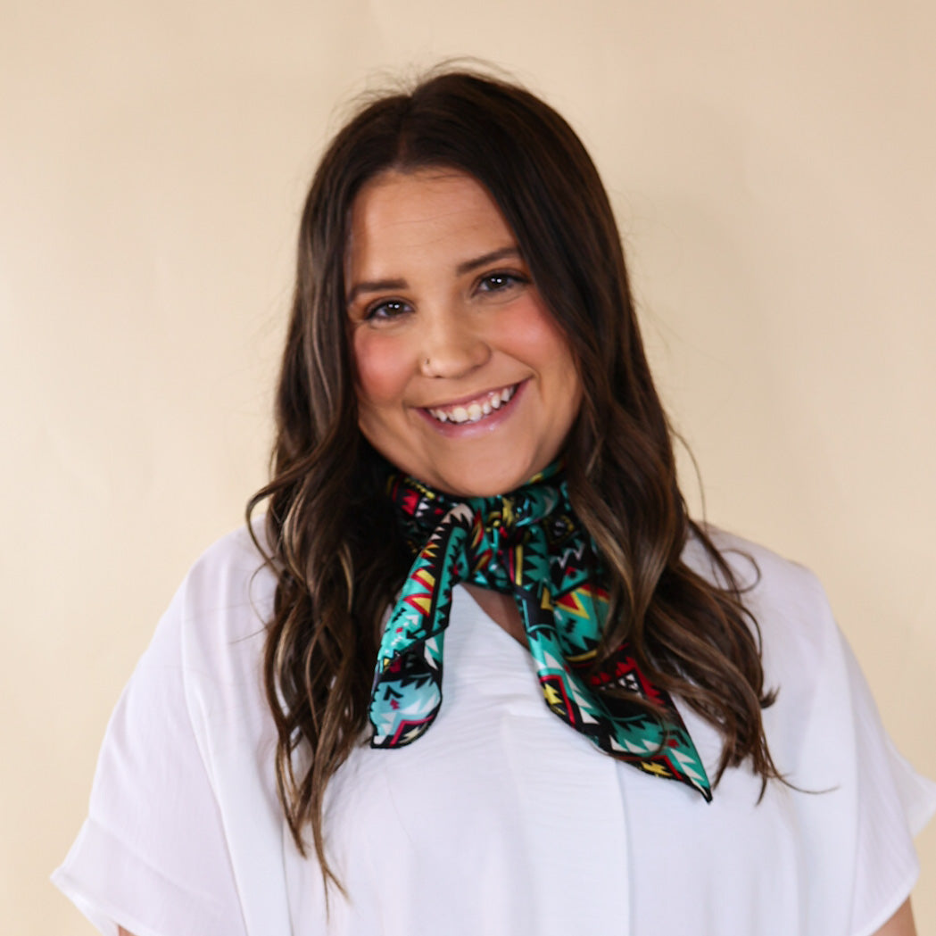 Brunette model wearing a white, drop shoulder top with multicolored print scarf with tied around her neck. Model is pictured in front of a beige background.