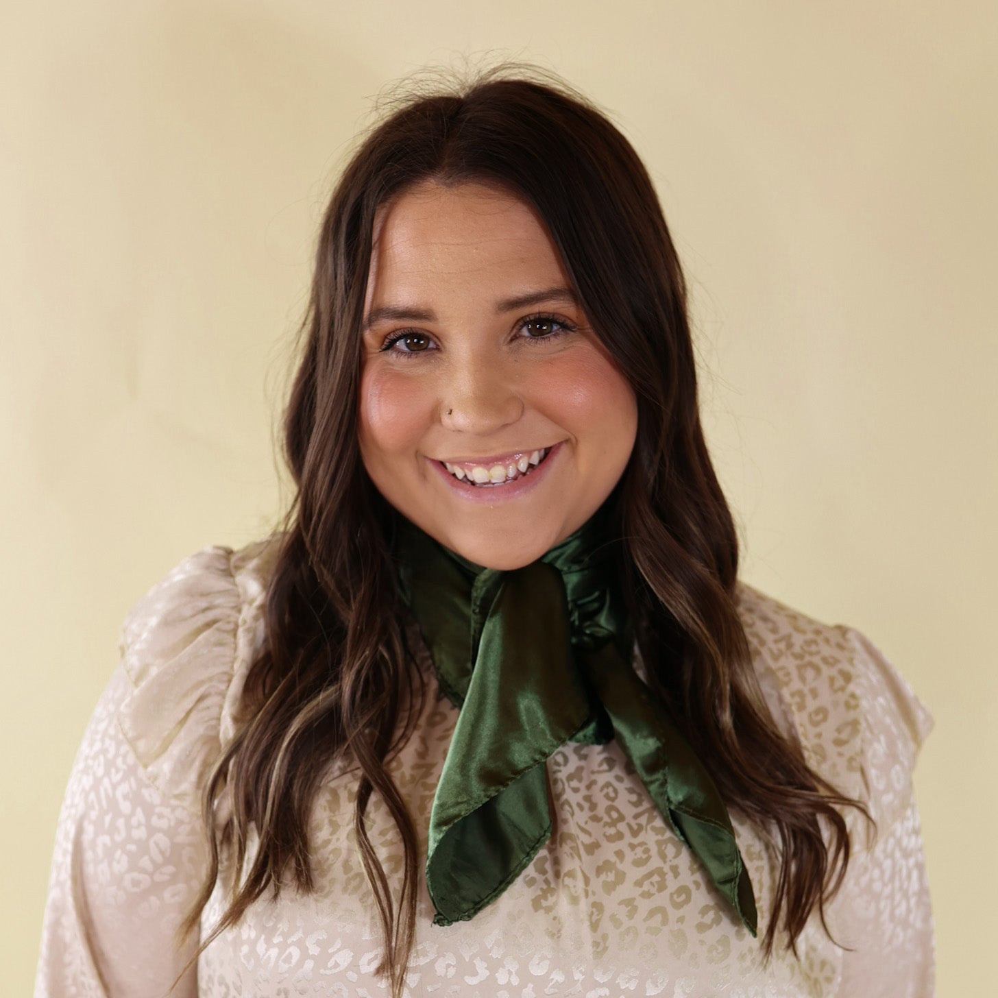 Brunette model wearing a long sleeve, iviry leopard print top with an olive green scarf tied around her neck. This model is pictured in front of a beige background. 