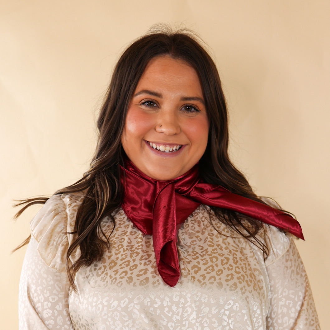 Brunette model is pictured wearing an invory, leopard print long sleeve top and a burgundy scarf tied around her neck. She is pictured in front of a beige background. 