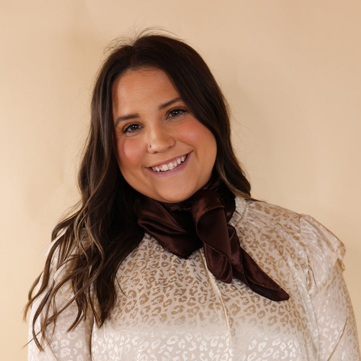 Brunette model is pictured wearing an invory, leopard print long sleeve top and a chocolate brown scarf tied around her neck. She is pictured in front of a beige background. 