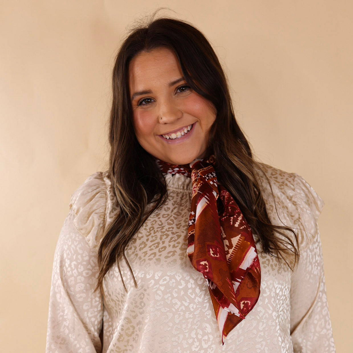Brunette model wearing an ivory, leopard print top with an red, brown, and white aztec print scarf tied around her neck. Model is pictured in front of a beige background. 