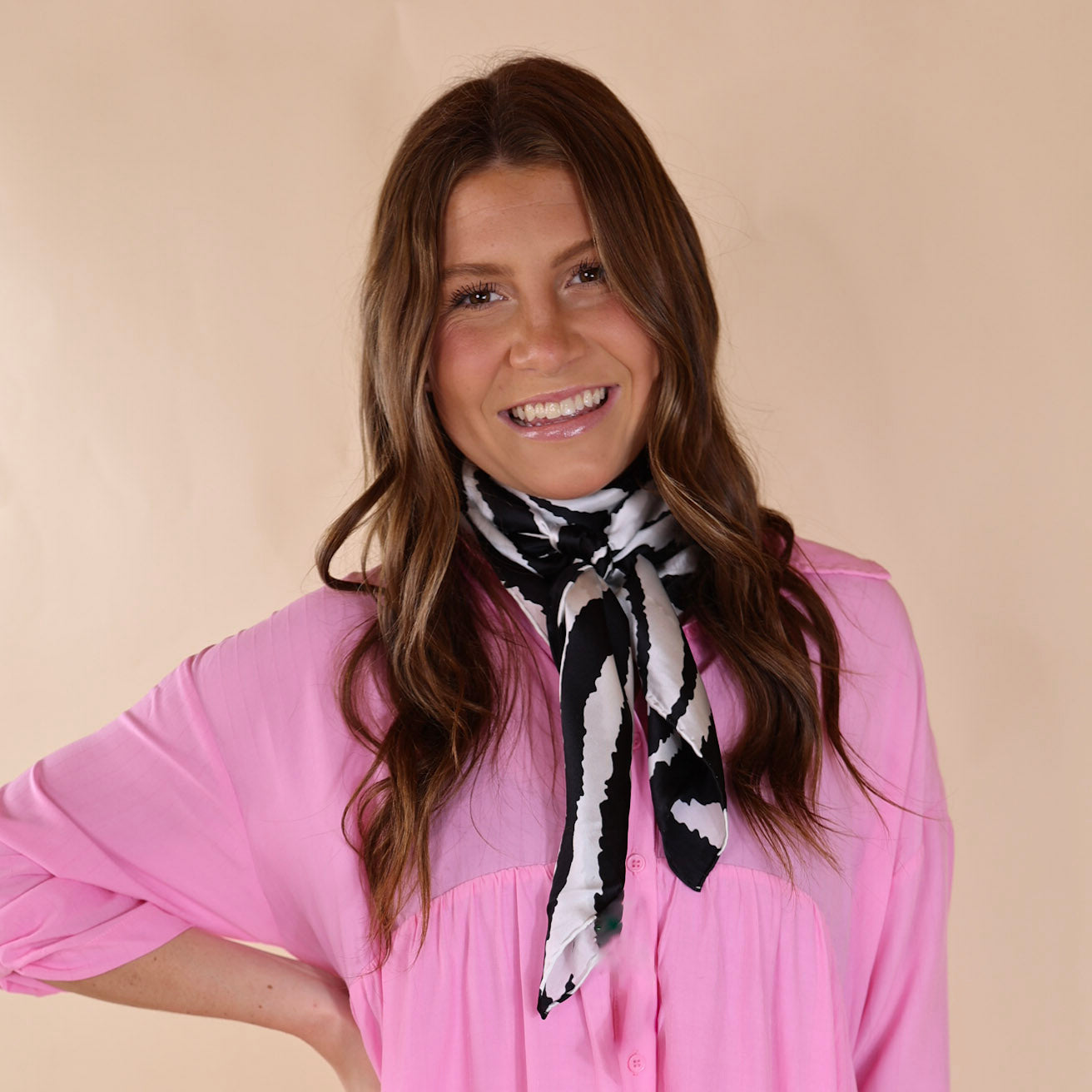 Brunette model wearing a pink, button up top with a black and white zebra print scarf tied around her neck. Model is pictured in front of a beige background. 